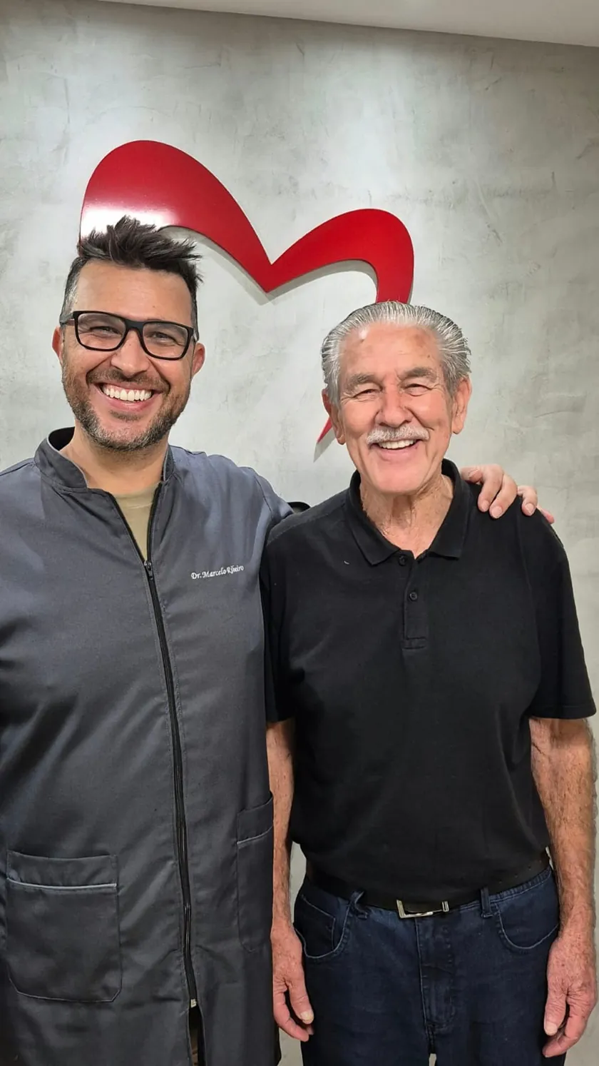 Paciente de camisa polo preta sorrindo abraçado ao Dr. Marcelo Ribeiro