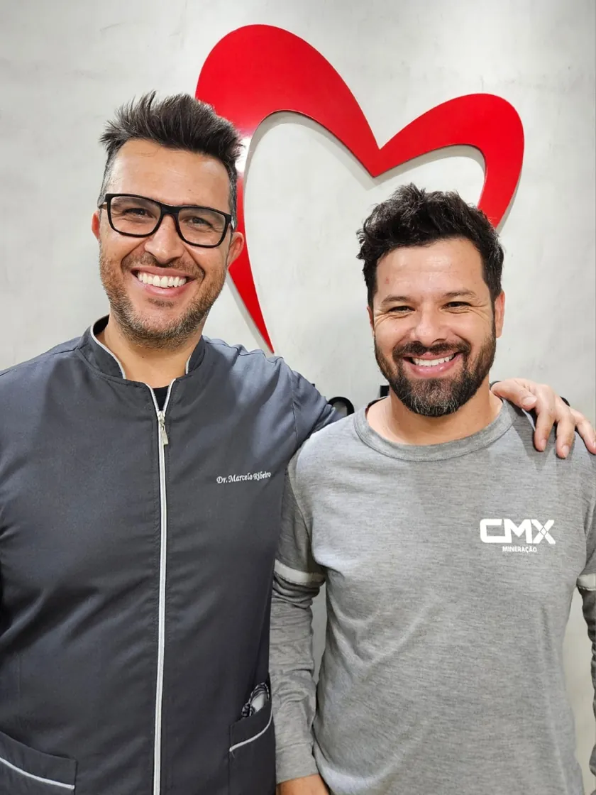Paciente de camiseta cinza sorrindo abraçado ao Dr. Marcelo Ribeiro